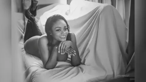 Smiling woman in minimal lingerie relaxing on a white bed in a bright, dreamy bedroom setting.