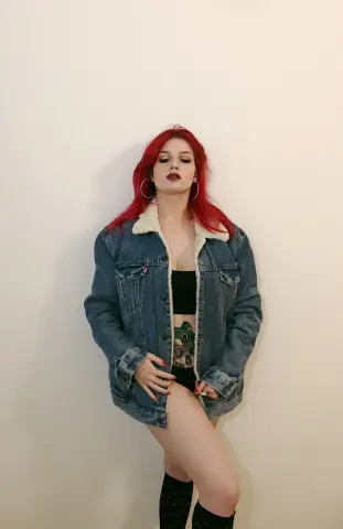 Red-haired woman in a denim jacket and black crop top posing against a white wall with a confident mood.