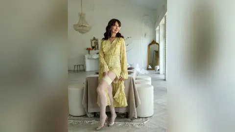 SophiaPanter smiles in a long yellow floral dress, sitting on a table in a bright room with her legs exposed and white stockings on.