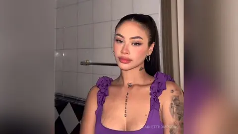 Young woman with tattoos in a purple ruffled top posing in a modern bathroom setting.