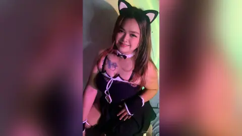 Smiling young woman in a black cat cosplay outfit with headband ears posing in soft colorful lighting.