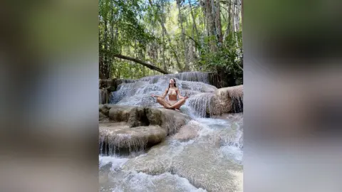 Serene woman in bikini practicing yoga meditation on a rocky waterfall in a lush tropical forest.