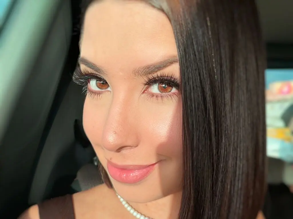 LuisanaVela smiles close-up, showing her long dark hair, brown eyes, and a white pearl necklace while sitting in a car.