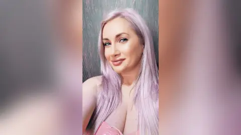 Smiling woman with long lavender hair wearing a pink top in a soft, bright indoor setting.