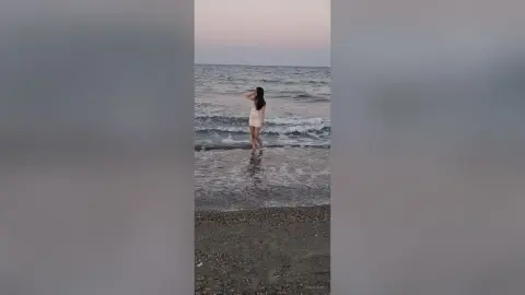 SaraTray stands in the shallow ocean waves at a beach, wearing a short white dress and looking out toward the horizon at dusk.