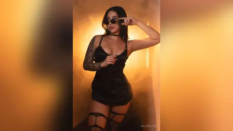 MilaWillow poses in a black slip dress and sunglasses against a warm, smoky background.
