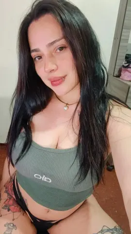 Smiling woman with long dark hair wearing an olive green ribbed tank top in a cozy indoor setting.