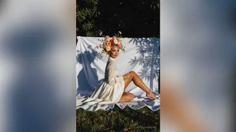 Elegant woman in a white flowing gown wearing a floral crown, sitting on a white sheet in a sunny garden setting.