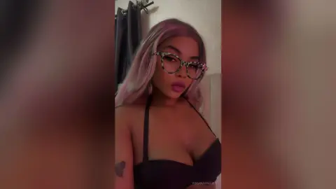 Young woman with pink hair and tortoiseshell glasses wearing a black bikini in a moody indoor setting.