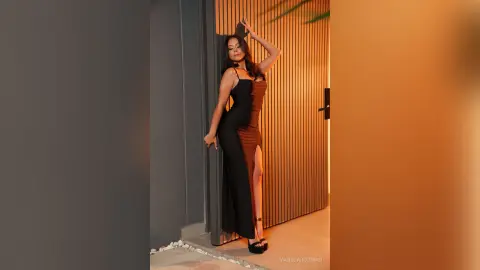 Elegant woman in a black evening gown posing by a modern slatted door in warm, moody lighting.
