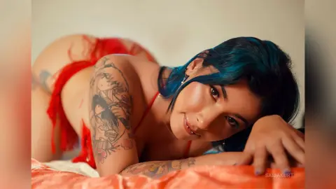 Tattooed woman with blue hair in red lingerie relaxing on an orange bed in a warm, intimate setting.
