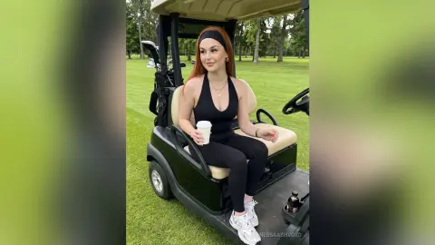 Redheaded woman in black athletic wear sitting in a golf cart on a sunny green golf course.