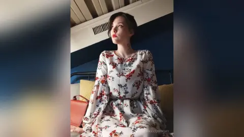 VikkiCominsky sits upright wearing a white long-sleeved floral print dress with a red flower pattern.