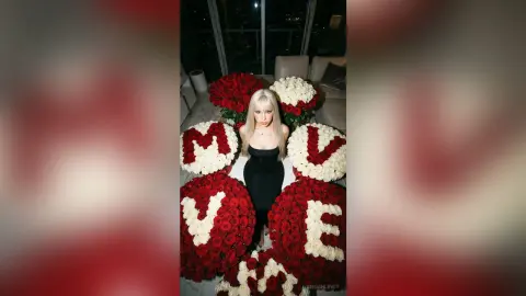 Blonde woman in a black dress sitting amidst large red and white flower arrangements spelling MOVE.