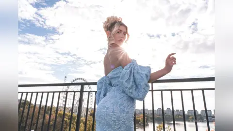 DaisyLin poses outdoors against a bright sky, wearing a light blue off-the-shoulder lace dress with blonde hair styled in a bun.