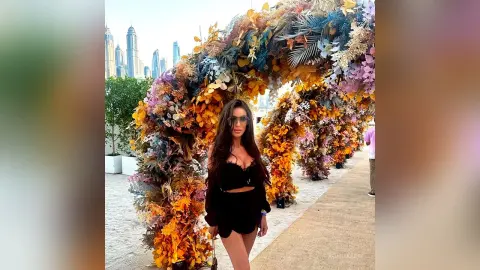 Woman in black outfit posing by a large floral archway with a city skyline background.