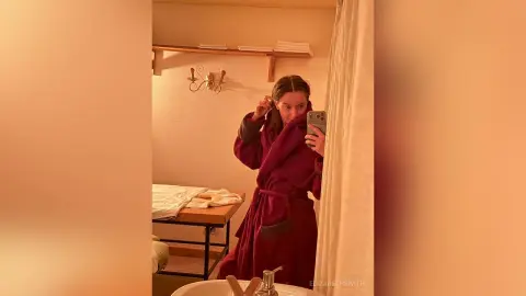 Young woman in a cozy burgundy bathrobe taking a mirror selfie in a warm, dimly lit bathroom setting.