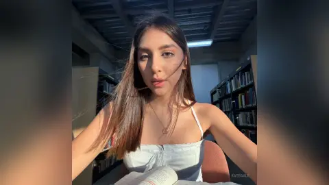 KateGale with long brown hair wearing a white camisole, sitting in a library setting with bookshelves behind her.