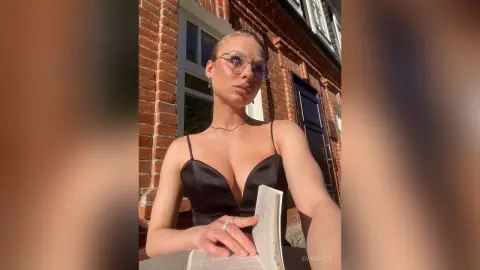 Stylish woman in a black satin camisole and glasses reading a book outside a classic brick building.