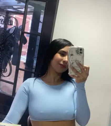 Young woman in a light blue long-sleeve crop top taking a casual mirror selfie indoors.