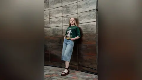 Young woman in a green graphic t-shirt and denim skirt leaning against a textured urban wall.