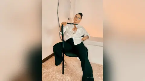 Stylish woman in white button-down shirt and black trousers sitting confidently in a minimalist modern room.