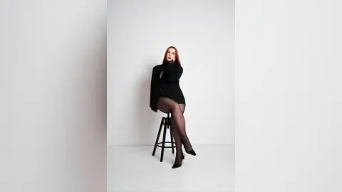 AryaSin poses sitting on a black stool, wearing a black blazer and sheer black tights with black heels against a white background.