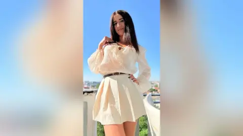 BellaLeen poses outdoors in a white long-sleeve blouse and a short cream skirt, smiling against a clear blue sky.