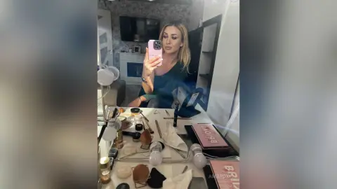Blonde woman in a dark green outfit taking a mirror selfie in a modern dressing room with makeup tools.