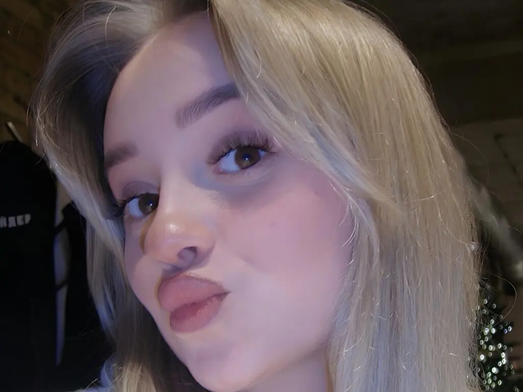 Blonde woman with long hair making a playful pouty face in a dimly lit indoor setting.