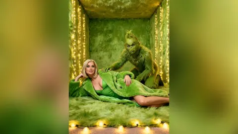 Blonde woman in a green dress and fur coat relaxing next to the Grinch in a festive green furry room.