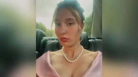Young woman in a pink dress and pearl necklace sitting in a car with a soft, natural look.