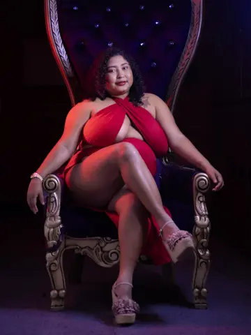 Curvy woman in a red plunging dress sitting on a regal purple velvet throne in moody lighting.
