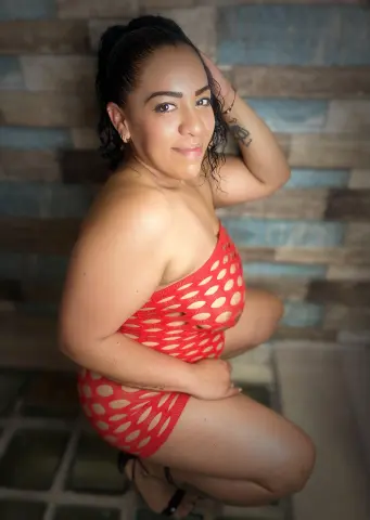Smiling woman in a red polka dot bodysuit posing against a rustic wood paneled wall.