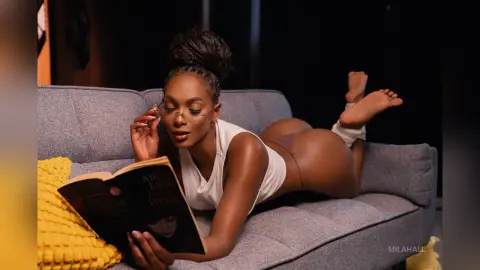 Young woman in white tank top reading a book while relaxing on a grey sofa in a cozy indoor setting.