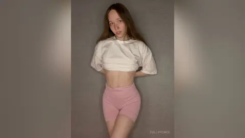 Young woman in a white crop top and pink yoga shorts posing against a grey wall in a casual mood.