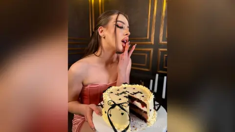 GracieCarter poses behind a large frosted cake, wearing a pink strapless top and touching her lips with one hand.