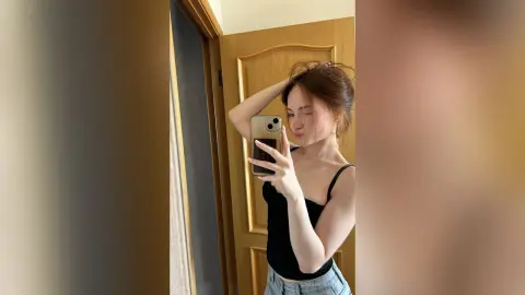 LauriRendleman poses in a black tank top and light blue denim shorts, holding her phone for a mirror selfie while touching her hair.