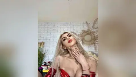Glamorous blonde woman in a red outfit posing with bold red lips in a bright, modern room.