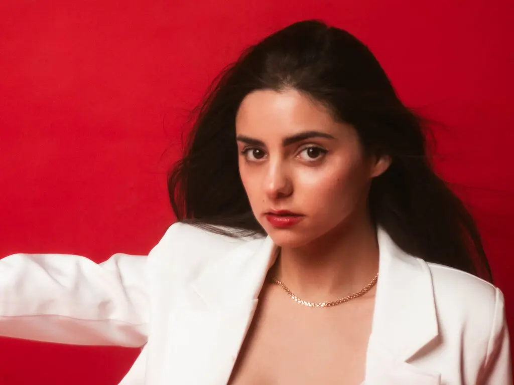 Elegant woman in a white blazer and gold necklace posing against a vibrant red background.