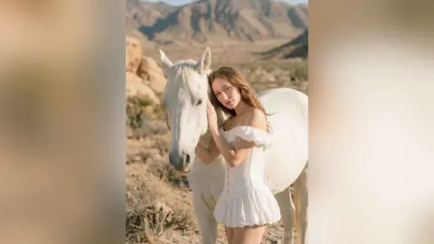 Dreamy portrait of a woman in a white sundress embracing a white horse in a desert mountain landscape.