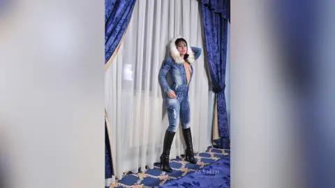 AndaCraig stands in front of white curtains wearing a blue denim jacket and matching denim jeans with black knee-high boots.
