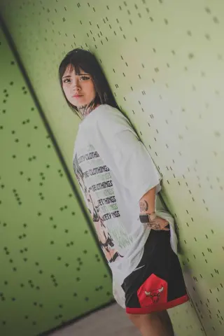 Young woman with long dark hair in an oversized white graphic t-shirt and black shorts leaning against a green wall.