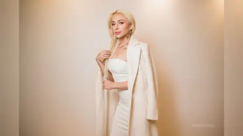 MiaRussell poses in a white dress and long white coat with her blonde hair flowing over her shoulder.