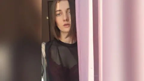 Young woman in a black sheer blouse posing by a pink curtain in soft, moody indoor lighting.