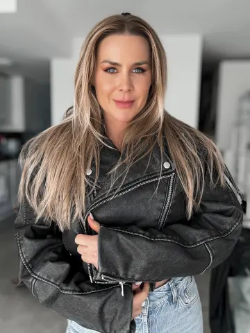 Smiling woman in a black leather jacket and light denim jeans posing in a bright indoor setting.