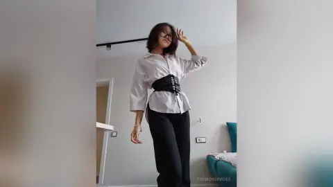 TishaOyervides poses in a white button-down shirt and black pants, wearing a black corset that cinches her waist.