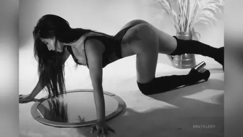 Black and white portrait of a woman in dark lingerie posing on a floor mirror in a minimalist studio setting.