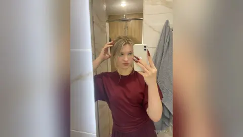 Young woman in a maroon t-shirt taking a casual mirror selfie in a modern bathroom.
