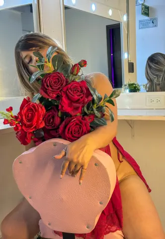 Woman in a red lace outfit holding a large bouquet of red roses in a bright dressing room.